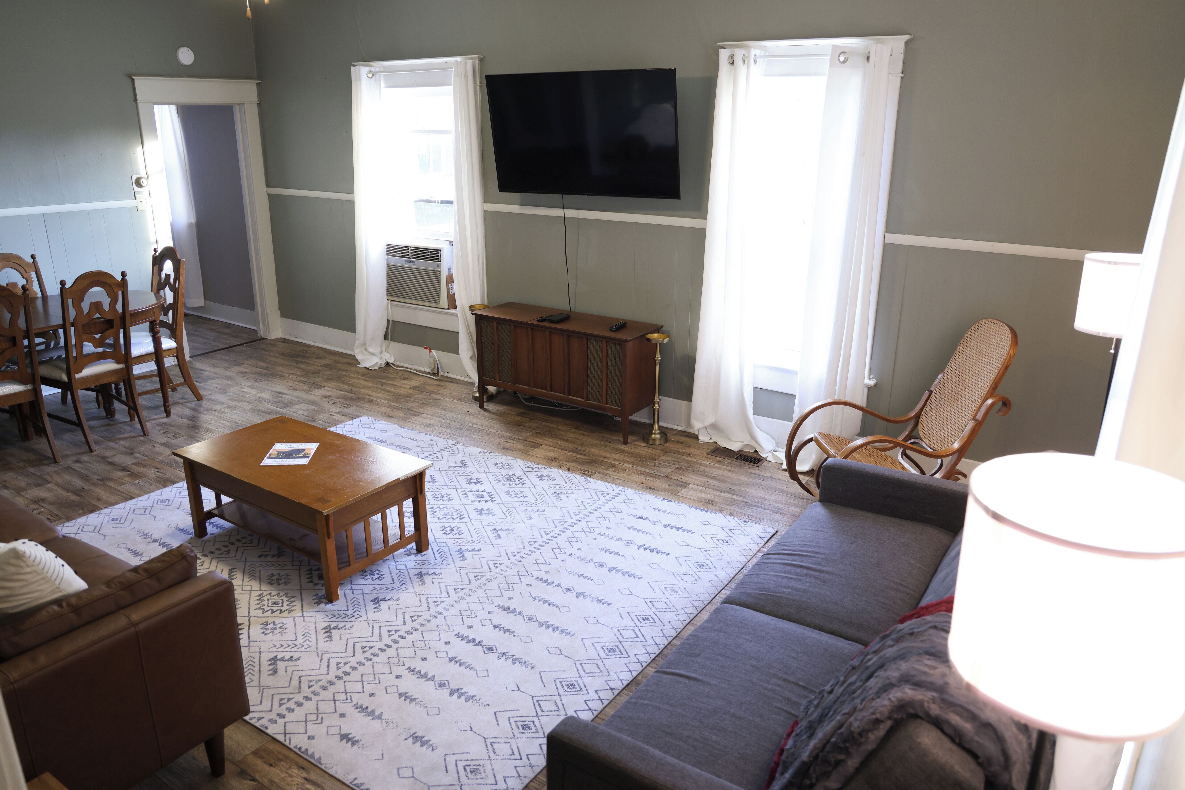 Una sala de estar con sofá, mesa de centro y sillas. Hay un televisor montado en la pared y una ventana con cortinas.