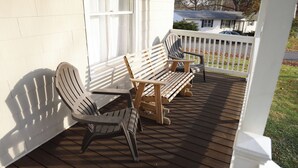 Una terraza de madera con dos sillas Adirondack y un banco, baranda blanca, y una casa al fondo.