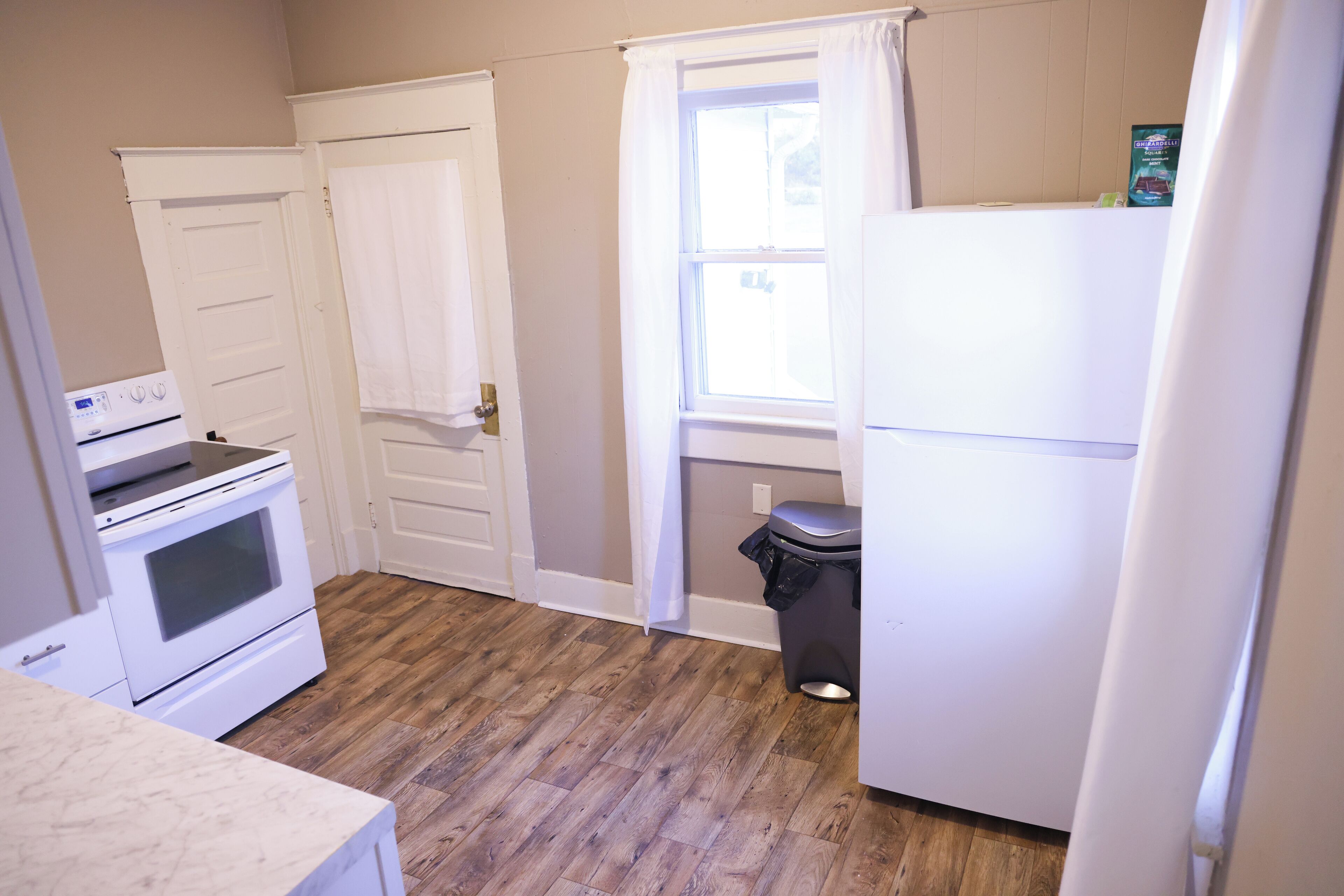 A kitchen with a white stove, oven, and refrigerator, wooden flooring, and a window with curtains.