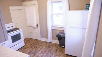 A kitchen with a white stove, oven, and refrigerator, wooden flooring, and a window with curtains.