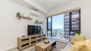 A modern living room with a flat-screen TV, wooden furniture, a large window with a city view, and a balcony.