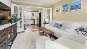A modern living room with a white sofa, a wooden coffee table, and a large plant. There is a kitchen in the background with white cabinets and stainless steel appliances. - Ogunquit River Outlook (Wells)