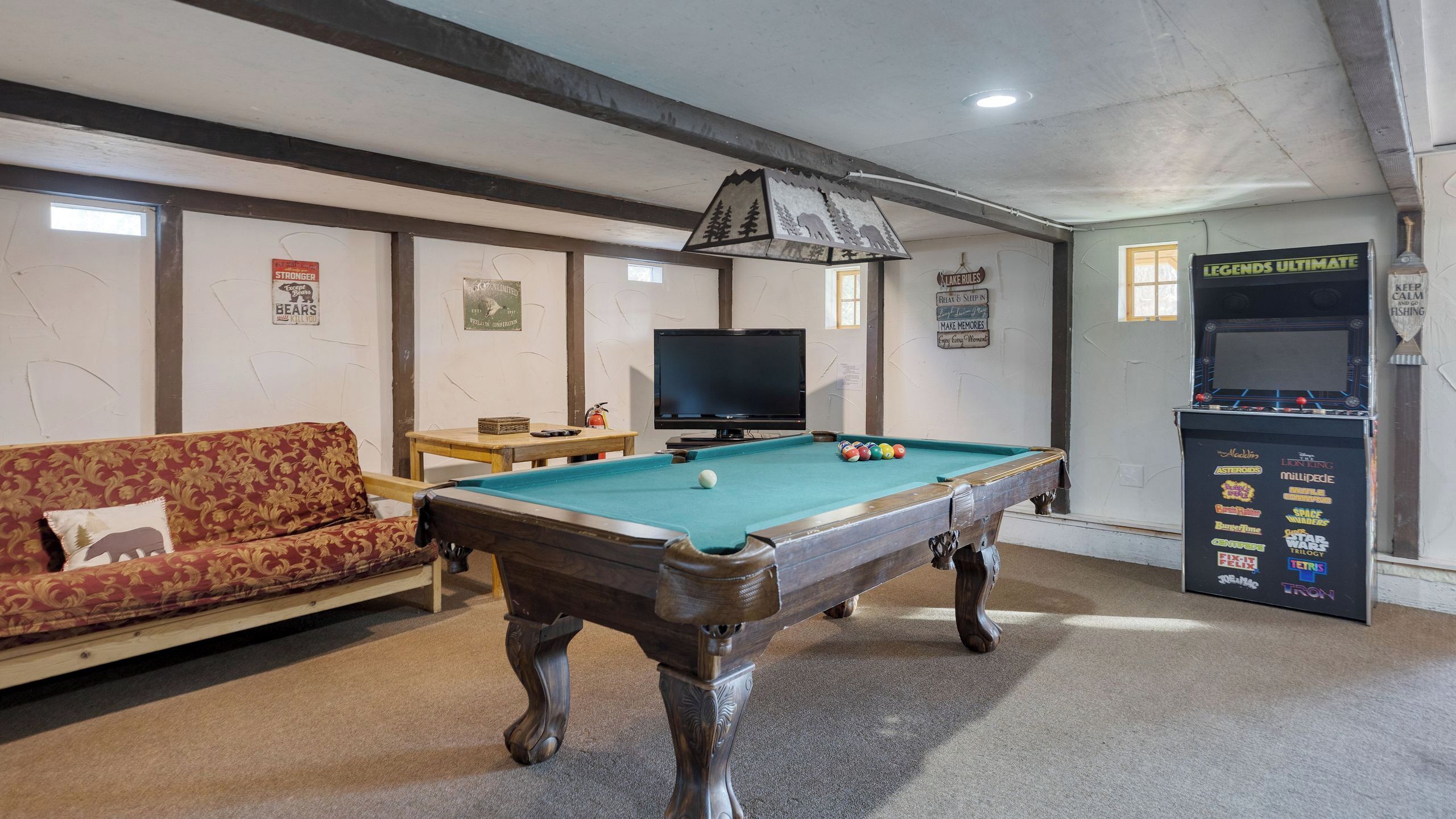 A pool table with balls, a sofa, a television, and a jukebox in a room with wooden beams and a sign that reads 'Lake Rules'.