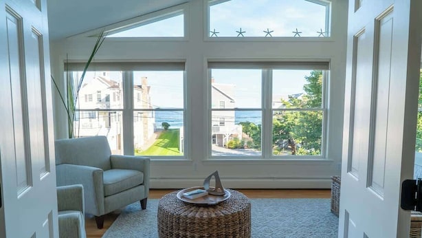 A room with a large bay window, a grey armchair, a woven ottoman, and a view of a residential area and water.