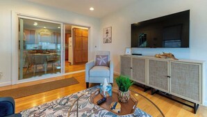A modern living room with a glass door leading to a dining area, a flat-screen TV mounted on the wall, and a cabinet with decorative items.