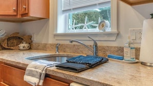 A kitchen with a granite countertop, a stainless steel sink, wooden cabinets, a window with blinds, and a view of trees outside.