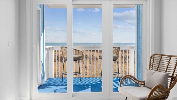 Habitación con vista a la playa a través de amplios ventanales correderos, una silla de mimbre y una terraza azul.