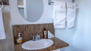 Ein Badezimmer mit einer Granit-Arbeitsplatte, einem weißen Waschbecken und einem Spiegel. Zwei weiße Handtücher hängen auf einem Halter und auf der Arbeitsplatte steht eine Flasche Handseife.