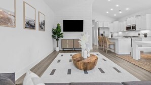 Ein modernes Wohnzimmer mit Couchtisch aus Holz, einem Flachbildfernseher und einer Küchenzeile mit weißen Schränken und einer Mikrowelle.