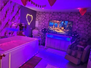 A room with a stone wall, a large flat-screen TV, a massage chair, and a bathtub. - Spa Suite la caverne Private terrace (Montpellier)