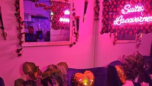 A room with a pink hue, featuring a sofa, a table with snacks, a tissue box, and a neon sign. - Spa Suite la caverne Private terrace (Montpellier)