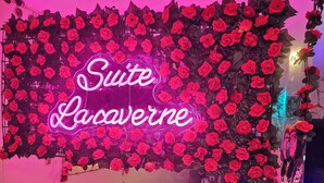 A room with a neon sign reading "Suite Lacaverne," surrounded by artificial red roses. There are dark blue chairs and a small table with items on it. - Spa Suite la caverne Private terrace (Montpellier)