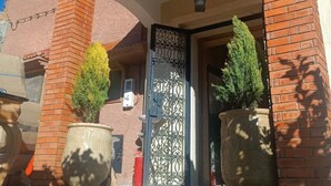 A building entrance with a wrought iron gate, two large potted plants, and a brick facade. - Gite Mrbie (Oum Rabia)