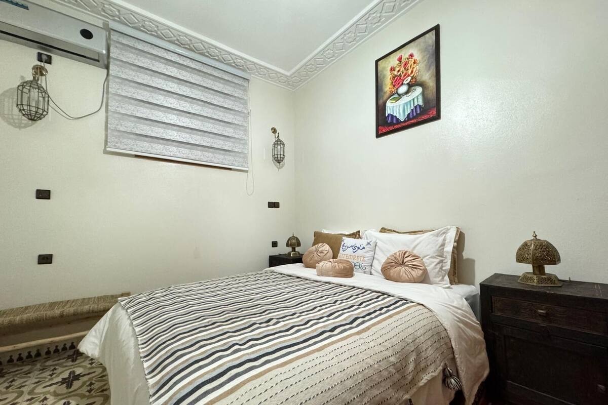 A bedroom with a bed, pillows, a nightstand, a lamp, a wall clock, and a painting.
