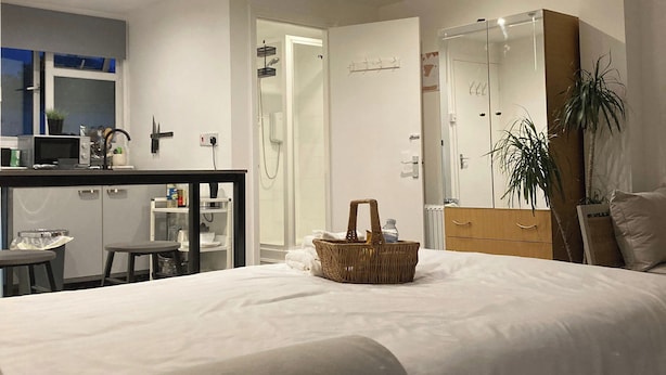 Ein modernes Hotelzimmer mit einem großen Bett, einer kleinen Küchenzeile, einem Badezimmer mit einer Glasdusche und einem Spiegelkorpus.