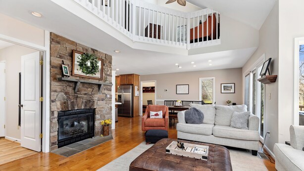 A spacious living room with a stone fireplace, a high ceiling with a fan, a grey sofa, and a brown leather ottoman.