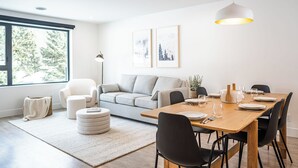 Ein modernes Wohnzimmer mit einem Holzespeisetisch, gedeckt für vier Personen, einem grauen Sofa und einem großen Fenster mit Blick auf Bäume.