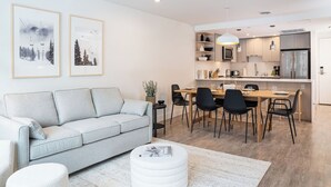 Ein modernes Wohnzimmer mit einer grauen Couch, einem Essbereich mit einem Holztisch und schwarzen Stühlen sowie einer Küche mit Edelstahlgeräten.