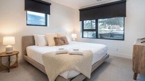 Ein modernes Schlafzimmer mit einem großen Bett, zwei Nachttischlampen, einem Holznachttisch und Blick auf eine verschneite Landschaft durch die Fenster.