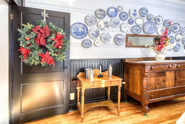 Une pièce avec une porte en bois sombre, une petite table en bois sur laquelle est posée une bougie, un miroir et un mur décoré d’assiettes bleues et blanches.