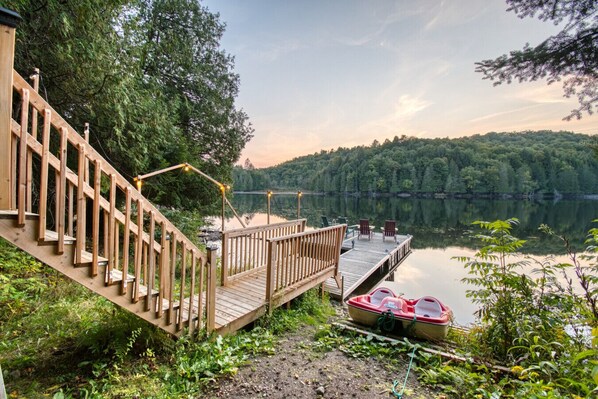 A wooden deck with chairs and a boat by a calm lake surrounded by trees.