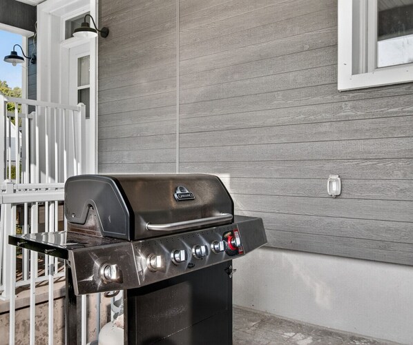 Una barbacoa de gas amb quatre cremalls i una prestatgeria lateral, col·locada davant d'una casa amb façana gris i adorns blancs.