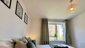 Un dormitorio con cama, almohadas, manta, lámpara colgante, ventana con cortinas y cuadros en la pared.