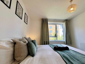A bedroom with a bed, pillows, a blanket, a hanging light fixture, a window with curtains, and framed pictures on the wall.