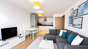 Una sala de estar moderna con un televisor de pantalla plana, un sofá gris con cojines azules y blancos, una mesa de centro blanca y una zona de comedor con una mesa y sillas blancas.
