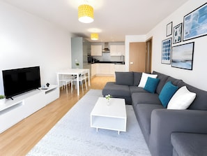 A modern living room with a flat-screen TV, a gray sofa with blue and white cushions, a white coffee table, and a dining area with a white table and chairs.