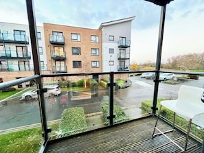 A balcony with a view of modern buildings, a parking area, and a wet ground.