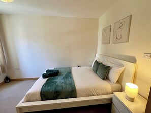 Un lit bien fait, avec une couverture verte et des oreillers, une lampe de chevet et deux œuvres d’art accrochées au mur.