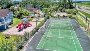 Een tennisbaan omringd door een omheind gebied met speeltoestellen en huizen op de achtergrond.