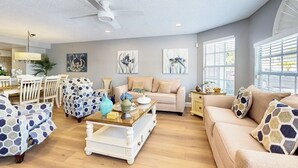 Een ruime woonkamer met een beige bank, gestreept fauteuils, een salontafel en een plafondventilator.