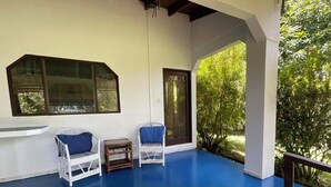 Eine überdachte Veranda mit zwei weißen Korbstühlen, blauen Kissen, einem kleinen Holztisch und einem großen Fenster.