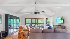 A spacious living room with a large grey sofa, a wooden coffee table, and a ceiling fan. There are large windows offering a view of the outdoors.
