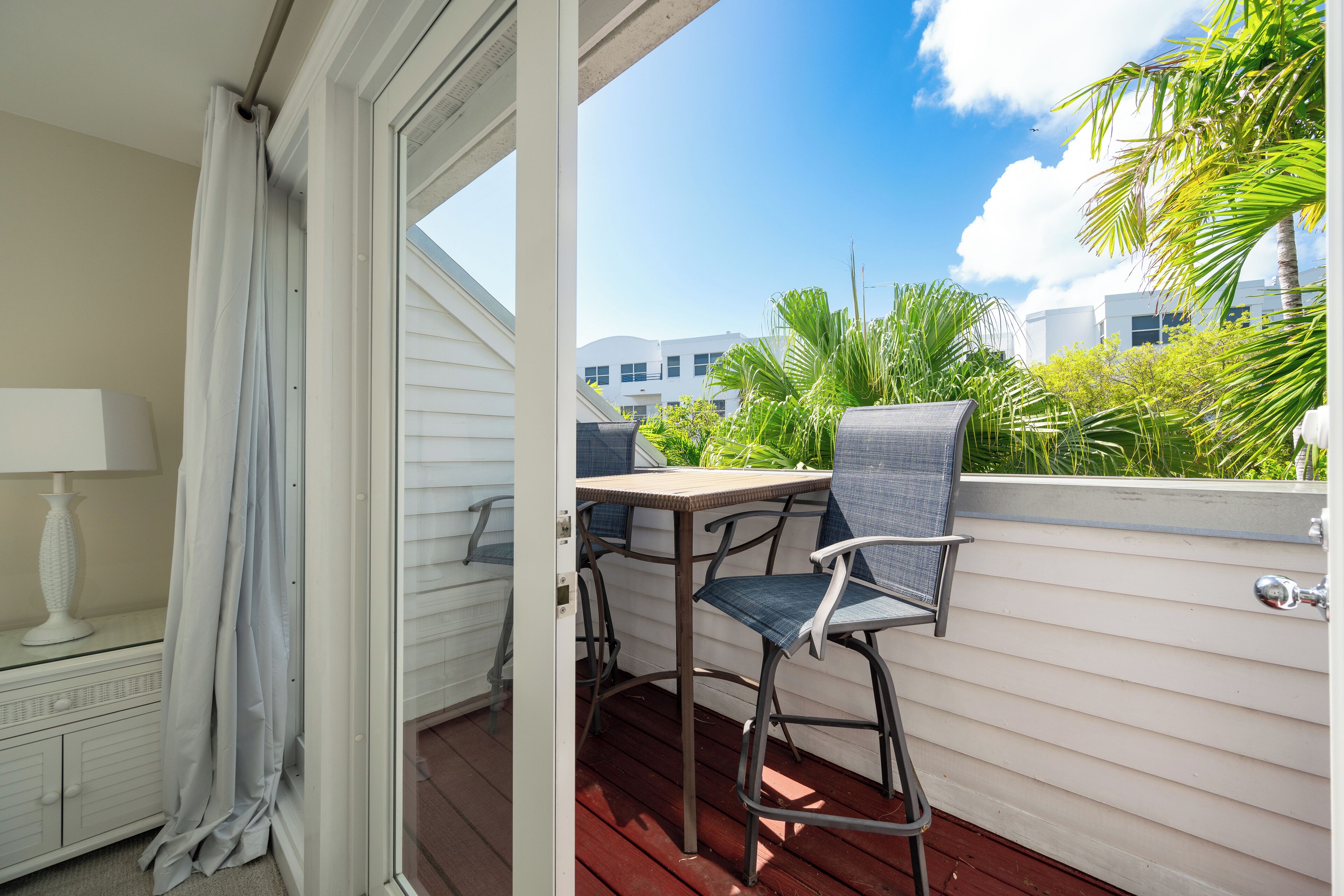 A balcony with a table and two chairs, a white cabinet with a lamp, and a view of buildings and greenery.