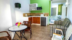 Un espacio habitable compacto con kitchenette, una zona de comedor con mesa y sillas, un sofá y una cama frente a una pared verde con cortinas.
