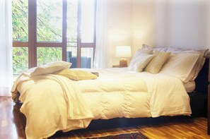 A neatly made bed with a white comforter and pillows, a bedside lamp, and a view of greenery through large windows. - QuickHome Villa "Il Tiglio" (Vigliano Biellese)
