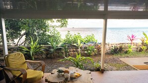 A covered patio with a view of the ocean, a wicker chair, a wooden table with drinks, and tropical plants. - Fare Taina by Tiare Lodge (Vai'anae)