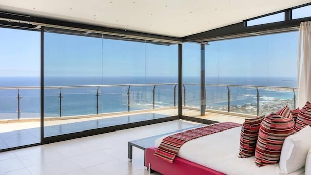 A modern bedroom with a large glass balcony, a bed with striped pillows, and a view of the ocean.