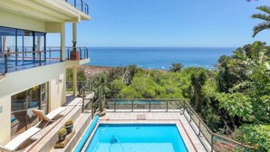 A pool with a view of the ocean, a balcony with lounge chairs, and lush greenery.