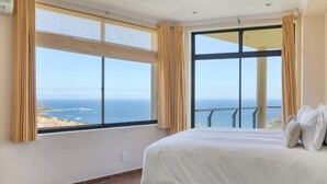 A bedroom with a large bed, two windows with curtains, and a view of the sea.