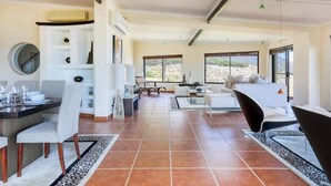 A spacious living area with a dining table, chairs, and a sofa. There are large windows providing natural light and a view of the outdoors.