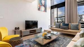 A modern living room with a large TV, comfortable seating, a glass coffee table, and a city view through large windows.