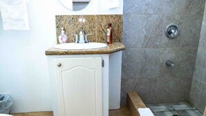 A bathroom with a granite countertop sink, a white cabinet, a mirror, and a shower area with a tiled floor. - The Hamlet Suites - Bright and spacious Studio Apartment 1 (Gros Islet)