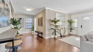 A modern living room with a wooden floor, a dining area with a bench, and a door with a decorative glass window. - Huge 8BR w/HTD Pool, Ensuites, Games, Near Beach (Palm Harbor)