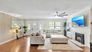A living room with a sectional sofa, a fireplace, a TV, and a ceiling fan. - Huge 8BR w/HTD Pool, Ensuites, Games, Near Beach (Palm Harbor)