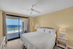 A bedroom with a large bed, a ceiling fan, a nightstand with a lamp, and a view of the ocean through a sliding door.