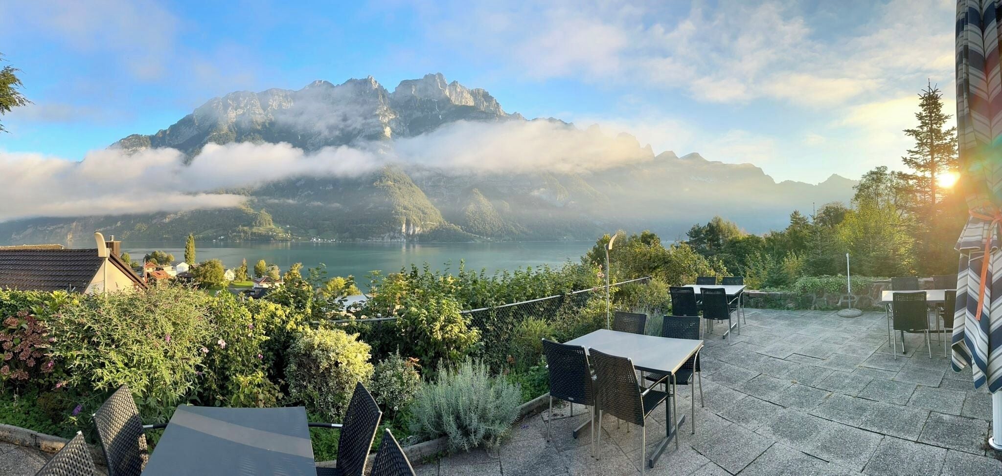Eine Terrasse mit Sitzgelegenheiten im Freien, mit Blick auf einen See und Berge, umgeben von viel Grün.